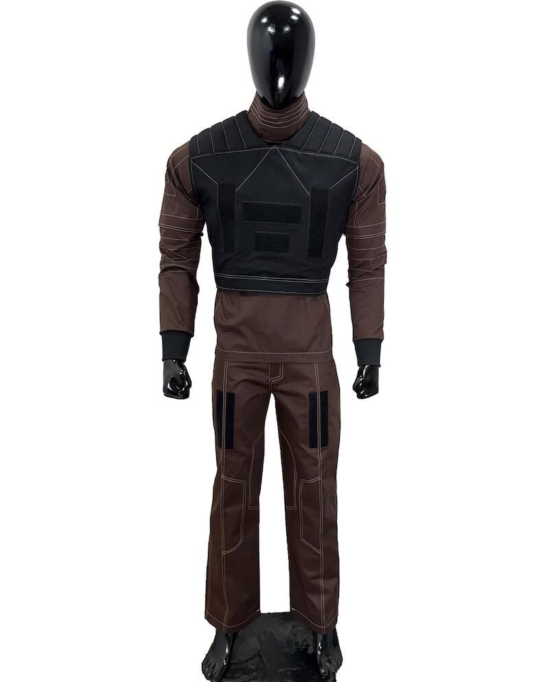 May include: A brown tactical outfit displayed on a mannequin. The outfit includes a long-sleeved shirt, pants, and a black vest. White stitching accents the seams. The mannequin has a black head and is standing on a black base.