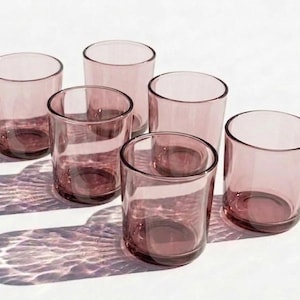 Peut inclure: Six verres à boire cylindriques transparents de couleur rose poussiéreux sont disposés sur une surface blanche. Les verres projettent des ombres qui reflètent la lumière. La verrerie convient à l'eau, aux jus ou aux cocktails.