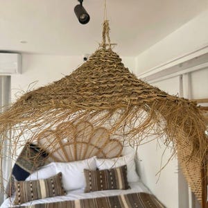 May include: A large, conical hanging light shade crafted from woven natural dried grass or straw. The shade features a textured, braided pattern with loose, frayed edges hanging down, suspended from a thin cord attached to a ceiling fixture.