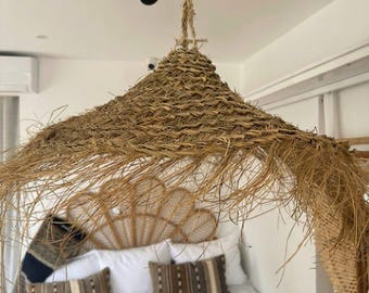 May include: A large, conical hanging light shade crafted from woven natural dried grass or straw. The shade features a textured, braided pattern with loose, frayed edges hanging down, suspended from a thin cord attached to a ceiling fixture.