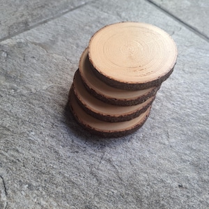 Puede incluir: Una pila de cinco posavasos redondos de madera. Cada posavasos está hecho de una sección transversal de una rama de árbol, mostrando la veta natural de la madera y los bordes de la corteza. Los posavasos son de color marrón claro con bordes marrones oscuros.