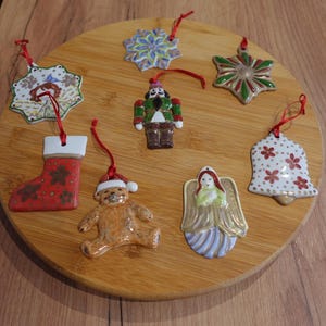Könnte beinhalten: Eine Sammlung von Weihnachtsbaumschmuck aus Keramik, darunter ein Strumpf, ein Teddybär, ein Engel, eine Glocke, ein Schneeflocke und ein Nussknacker. Jedes Ornament ist handbemalt mit festlichen Farben und Designs und hat ein rotes Band zum Aufhängen. Die Ornamente sind auf einer Holzoberfläche angeordnet.