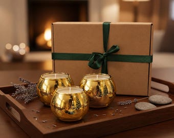 May include: Three gold-colored spherical candle holders with white wax, arranged on a dark wooden tray alongside a brown cardboard gift box tied with a dark green ribbon, dried lavender sprigs, and two smooth gray stones.