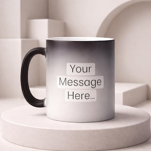 Può includere: Una tazza bianca che cambia colore con un manico nero. La tazza ha il testo "Your Message Here..." stampato sul davanti. La tazza è su un piedistallo bianco.