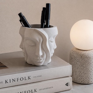 May include: A white, sculpted face pen holder with multiple faces, holding black pens and markers. The holder sits on a stack of books with the title "The Kinfolk". A spherical lamp is in the background.