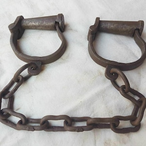 Puede incluir: Grilletes antiguos de hierro con una cadena de conexión. Los grilletes tienen un diseño en forma de U con un asa y están conectados por una serie de eslabones de cadena de hierro. El metal tiene un aspecto oscuro y envejecido.