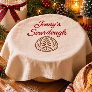 May include: A beige fabric proofing cover featuring the embroidered red text Jenny's Sourdough above a brown stitched wheat stalk design. The circular cloth is draped over a round bowl, showcasing a textured weave and clean edges.