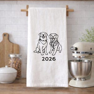 Puede incluir: Un paño de cocina blanco con una ilustración de líneas negras de dos perros sentados uno al lado del otro, uno con un pañuelo y el otro con un arnés. El número 2026 está impreso en texto negro justo debajo de la ilustración de los perros.