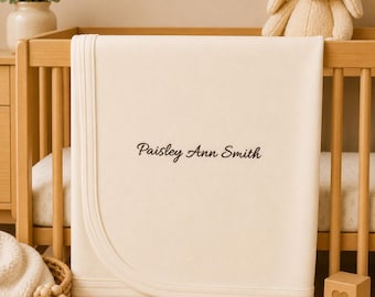 May include: A cream-coloured baby blanket featuring the embroidered name Paisley Ann Smith in dark brown script lettering. The blanket has a smooth, solid-coloured fabric surface with a finished stitched border, draped over the side of a wooden crib.