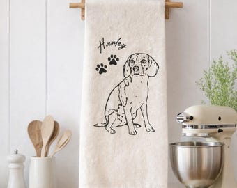 May include: A cream-coloured kitchen towel hanging on a wooden rack, featuring a black line-art illustration of a dog, two paw prints, and the name Harley written in a script font above the dog's head on the centre of the fabric.
