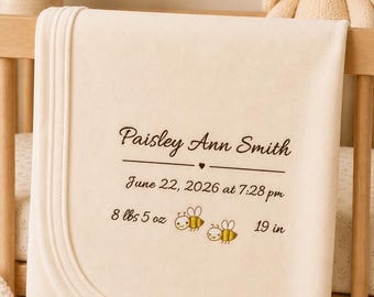 May include: A cream-coloured baby blanket featuring dark brown embroidered text that reads Paisley Ann Smith, June 22, 2026 at 7:28 pm, 8 lbs 5 oz, and 19 in, accompanied by two small embroidered bees with yellow and brown stripes.