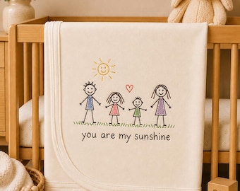 May include: A cream-coloured baby blanket featuring an embroidered stick figure family of four, a yellow sun, a red heart, and green grass. The text "you are my sunshine" is embroidered in black below the figures on the soft fabric surface.