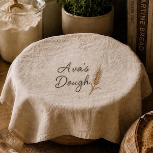 May include: A round, beige linen dough cover featuring dark brown embroidered text that reads Ava's Dough, accompanied by a small embroidered wheat stalk design. The fabric has a textured, natural weave and drapes over a circular bowl.