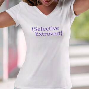 May include: White V-neck t-shirt with the text "{Selective Extrovert}" in purple. The shirt is made of a soft material and has short sleeves. The design is centered on the chest.