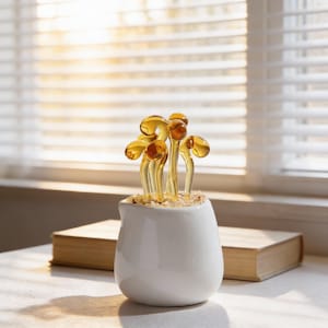 May include: A small white ceramic pitcher filled with amber-coloured glass mushrooms. The mushrooms have a detailed, realistic design. The pitcher sits on a white surface with a closed book in the background, near a window with blinds.