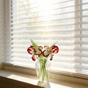 Könnte beinhalten: Eine klare Glasvase mit drei Glasblumen mit roten und klaren Blütenblättern sowie grünen Stielen und Blättern. Die Vase steht auf einer Fensterbank vor einem Fenster mit weißen Jalousien.