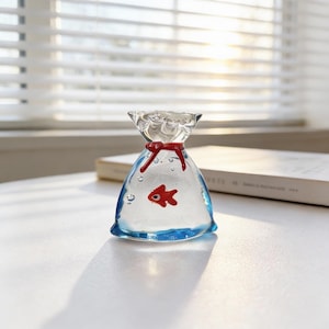 Puede incluir: Una escultura de vidrio transparente de un pez dorado en una bolsa. La bolsa está atada con una cinta roja y tiene una base azul. Un pequeño pez rojo es visible dentro de la bolsa. La escultura está sobre una superficie blanca.