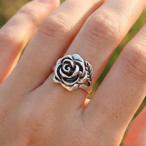 Könnte beinhalten: Ein silberner Ring mit einem Rosenmotiv. Der Ring zeigt eine detaillierte Rose mit komplizierten Blütenblattdetails und zwei kleinen Blättern an der Seite. Der Ring wird an einem Finger getragen.
