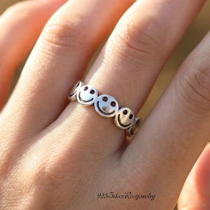 May include: A silver ring featuring a repeating pattern of smiley faces. The ring is worn on a finger, and the design is visible. The text "925SilverRootjewelry" is at the bottom of the image.