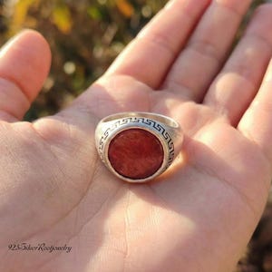 Op de afbeelding: Een zilveren ring met een rode steen in het midden, vastgehouden in een open hand. De ring heeft een Grieks sleutelontwerp rond de steen. De ring is een zegelring, met een ronde rode steen.