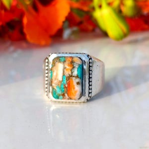 May include: A silver ring with a rectangular, multi-colored stone. The stone features a mosaic of turquoise, orange, and white hues. The ring has a simple, classic design with a beaded border around the stone.