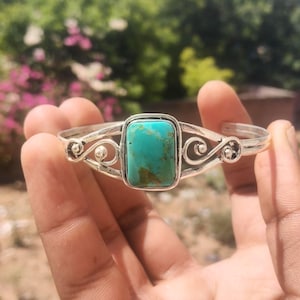 May include: A silver cuff bracelet featuring a rectangular turquoise stone. The bracelet has an open design with decorative swirls and small ball accents on either side of the stone. The turquoise stone is a vibrant blue-green color.
