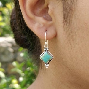 May include: A silver dangle earring featuring a square-cut turquoise stone set in a metal frame with small decorative silver beads at the top and bottom points. The earring hangs from a simple silver hook wire that passes through the earlobe.