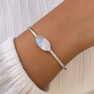 May include: A silver cuff bracelet featuring a central oval-shaped moonstone with a smooth, polished finish. The metal band is thin and rounded, connecting to the bezel setting that holds the iridescent, translucent gemstone in place.