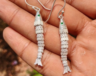May include: A pair of silver fish-shaped dangle earrings featuring textured scales and a small green stone set as an eye. The earrings hang from curved silver ear wires and have a segmented body design that mimics the appearance of a fish.