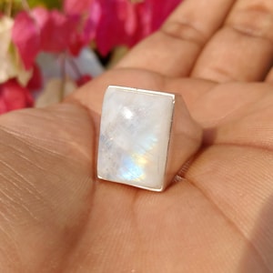 May include: A silver ring featuring a large, rectangular, translucent moonstone cabochon. The stone displays a milky white appearance with subtle iridescent blue and yellow flashes across its smooth, polished surface, set within a plain metal band.