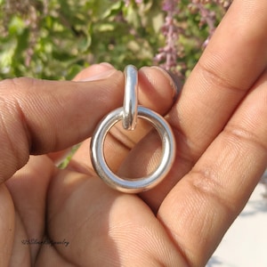 Peut inclure: Un pendentif argenté avec un design circulaire et une boucle de connexion. Le bijou est tenu dans une main, avec un arrière-plan flou de feuillage vert et violet. Le pendentif a un design simple et élégant.