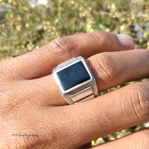 Könnte beinhalten: Ein silberner Ring mit einem rechteckigen schwarzen Stein in einem detaillierten Band. Der Ring wird an einem Finger getragen und zeigt sein Design und seine Handwerkskunst. Das Band des Rings weist eingravierte Muster auf.