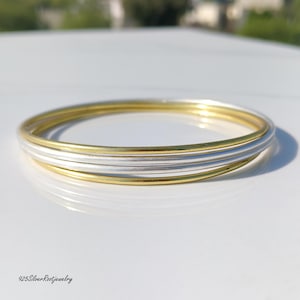 Puede incluir: Una pila de tres pulseras finas, dos doradas y una plateada, apiladas sobre una superficie blanca. Las pulseras son lisas y brillantes, reflejando la luz. La joyería es sencilla y elegante, adecuada para el uso diario.