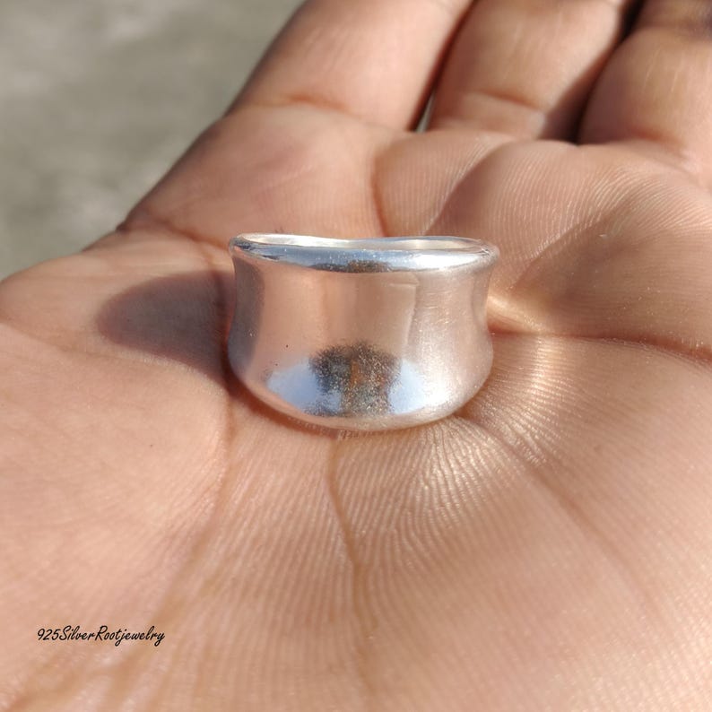 May include: A silver ring with a rounded, flared design. The ring is smooth and reflective, with a polished finish. The ring is shown in a close-up shot, highlighting its shape and texture. The ring is a piece of jewelry.