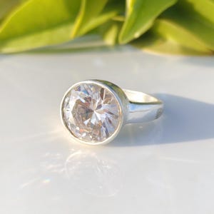 May include: A silver ring featuring a large, round, clear gemstone. The gemstone is faceted, reflecting light and creating a sparkling effect. The ring band is simple and polished, complementing the prominent stone. The ring is set against a white surface with green foliage in the background.