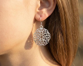 May include: A silver filigree earring is shown hanging from an ear. The earring has a circular design with an open, floral pattern. The earring is suspended from a simple hook and wire. The background is blurred, with a hint of sunlight.