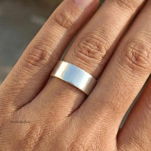 May include: A silver band ring with a flat, wide design. The ring has a smooth, shiny surface, reflecting light. The ring is worn on a finger. The ring is a simple, classic piece of jewellery.