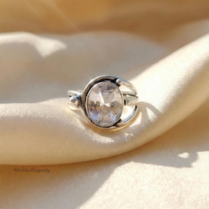 Könnte beinhalten: Ein silberner Ring mit einem ovalen, klaren Edelstein. Der Ring hat ein dekoratives Band mit geschwungenen Akzenten. Der Text "925-SilverRootjewelry" ist sichtbar. Der Ring liegt auf einem cremefarbenen Stoff.
