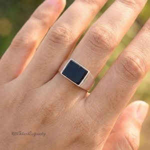 Könnte beinhalten: Ein silberner Ring mit einem rechteckigen schwarzen Onyxstein. Der Ring hat ein poliertes Silberband. Der Schmuck wird auf einer Hand gezeigt.