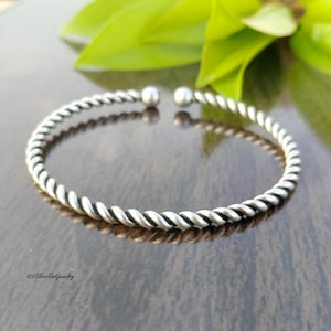 May include: A silver-colored, open cuff bracelet with a twisted rope design. The bracelet has a smooth, rounded finish and is displayed on a reflective surface. The text "925 Silver Root Jewelry" is visible.