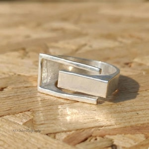 Puede incluir: Un anillo plateado con un diseño geométrico. El anillo presenta una barra rectangular conectada a una banda curva. El anillo de metal brillante está sobre una superficie de madera.