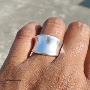 May include: A wide, polished silver ring with a smooth, curved design. The ring is shown on a finger, reflecting the sunlight. The jewelry piece has a minimalist style and a bright, shiny finish. The ring is a statement piece.