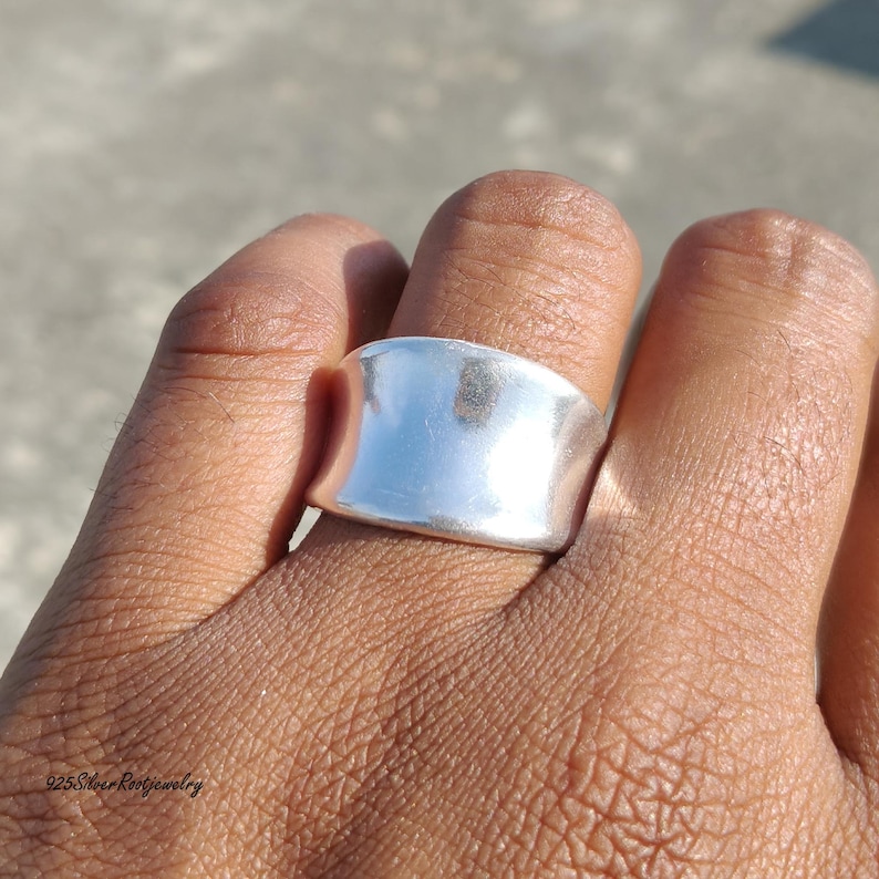 May include: A wide, polished silver ring with a smooth, curved design. The ring is shown on a finger, reflecting the sunlight. The jewelry piece has a minimalist style and a bright, shiny finish. The ring is a statement piece.
