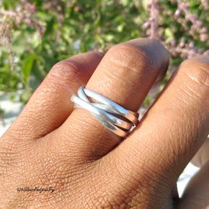 Könnte beinhalten: Ein silberner Ring mit einem dreifachen Banddesign. Die Bänder sind miteinander verwoben und erzeugen einen einzigartigen und modernen Look. Der Ring wird an einem Finger getragen und zeigt seine polierte Oberfläche. Der Ring ist ein Schmuckstück.