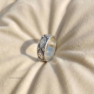 May include: A silver ring with an intricate pattern of diamonds, dots, and lines. The ring is made of a shiny metal and has a detailed design around the entire band. The ring is displayed on a soft, beige surface.