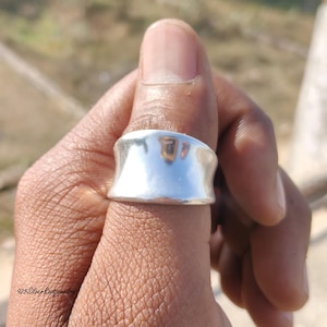 May include: A wide, polished silver ring with a curved design. The ring is worn on a finger, reflecting the surrounding environment. The image is taken in natural light, highlighting the ring's smooth surface. The text "925 Silver Root Jewelry" is visible at the bottom left.