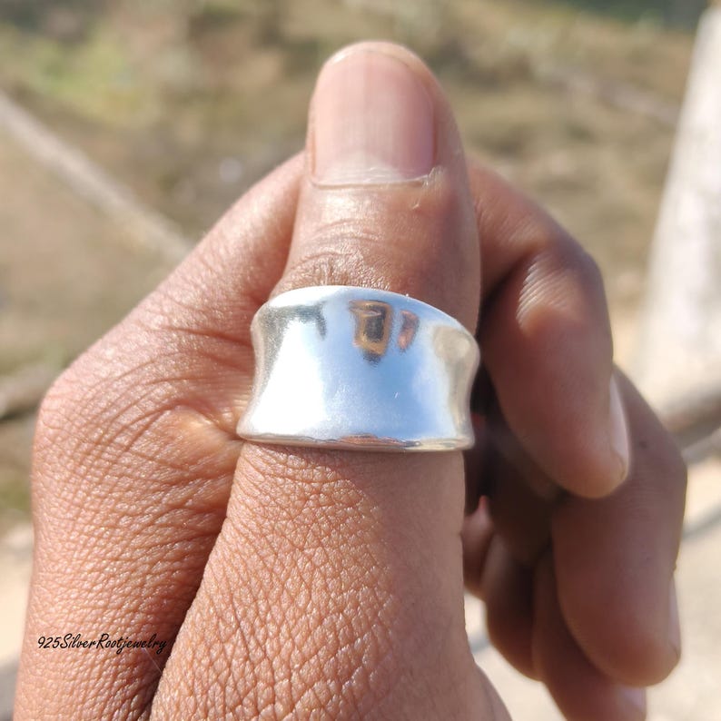 May include: A wide, polished silver ring with a curved design. The ring is worn on a finger, reflecting the surrounding environment. The image is taken in natural light, highlighting the ring's smooth surface. The text "925 Silver Root Jewelry" is visible at the bottom left.