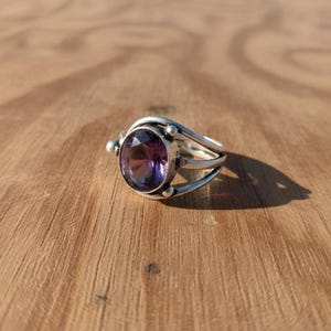 May include: A silver ring with a large, oval-cut purple gemstone. The ring has a split band design with small decorative beads on the sides. The gemstone is a deep purple color, and the ring is set against a wooden surface.