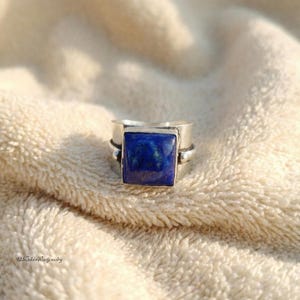 Puede incluir: Un anillo de plata con una gran piedra cuadrada de lapislázuli azul oscuro. El anillo tiene una banda gruesa y un diseño simple y moderno. La piedra está engastada en un bisel, sobre una superficie texturizada de color crema.