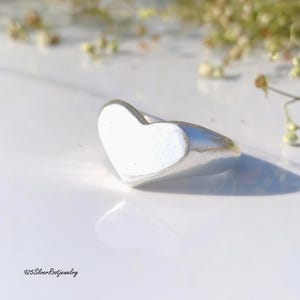 May include: A silver ring with a heart-shaped design. The ring is made of polished metal and has a smooth, reflective surface. The heart is white and the ring is on a white surface. The text "925SilverRootJewelry" is at the bottom left.